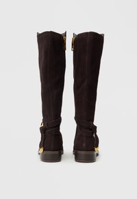 Čizme do koljena, tamnosmeđe odsušene kože s teksturiranom površinom, bočnim patentnim zatvaračima i dekorativnim trakama s zlatnim detaljima. Ravni drveni potplati.
