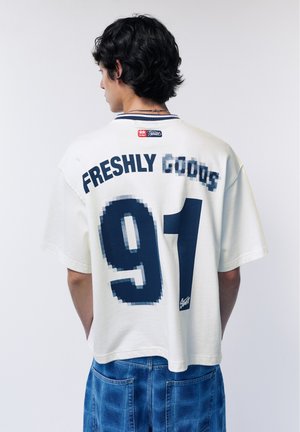 Persona con capelli corti e neri indossa una camicia bianca oversize con la scritta "FRESHLY GOODS" e il grande numero "91" sul retro, abbinata a jeans blu.