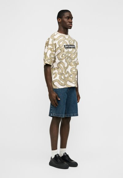 Ανδρικό κοντομάνικο μπλουζάκι σε κρεμ απόχρωση με χρυσό αφηρημένο σχέδιο και μαύρο λογότυπο. Συνδυάζεται με μπλε τζιν σορτς και μαύρα αθλητικά παπούτσια.
