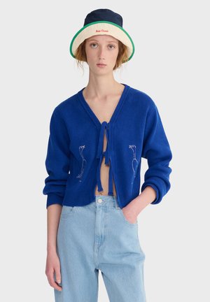 Jonge vrouw met een blauwe cardigan die voorgebonden is, lichtblauwe spijkerbroek en een tweekleurige bucket hat tegen een effen achtergrond.