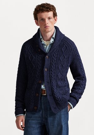 Man wearing a dark blue chunky cable-knit cardigan over a light blue button-up shirt and dark jeans, standing with one hand in pocket.