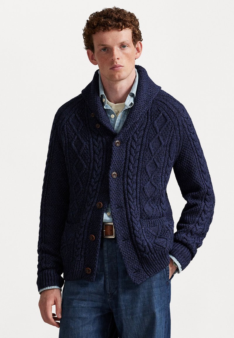 Hombre vistiendo un cárdigan de punto grueso en azul oscuro sobre una camisa de botones en azul claro y jeans oscuros, de pie con una mano en el bolsillo.