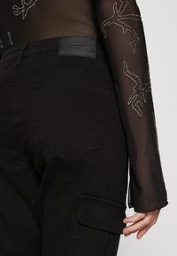 Nära bild av svarta cargobyxor med bakficka och märkesläderlapp, kombinerat med en genomskinlig långärmad topp med rhinestones-mönster.