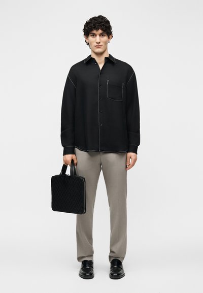 Άνδρας στέκεται κρατώντας μαύρη υφαντή τσάντα, φοράει μαύρο πουκάμισο με κουμπιά και λευκή ραφή, μπεζ παντελόνι και μαύρα επίσημα παπούτσια.