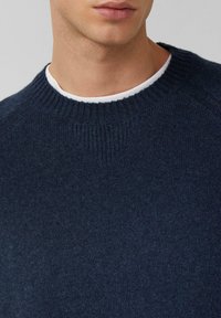 Zbliżenie na mężczyznę w granatowym swetrze dzianinowym, noszącym biały t-shirt z okrągłym dekoltem, pokazujące dekolt i dolną część twarzy.