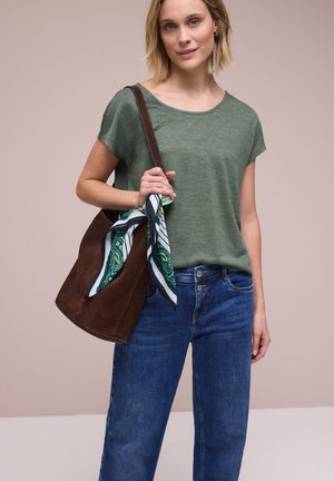 Donna che indossa una maglietta verde a maniche corte e jeans blu, con una borsa a tracolla marrone e una sciarpa con motivi verdi e bianchi.