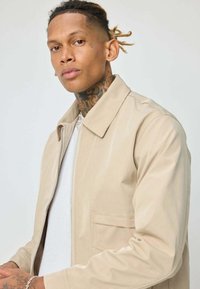 Jeune homme avec des tatouages sur le cou et la main, portant une veste beige sur une chemise blanche, se tenant devant un fond clair uni.