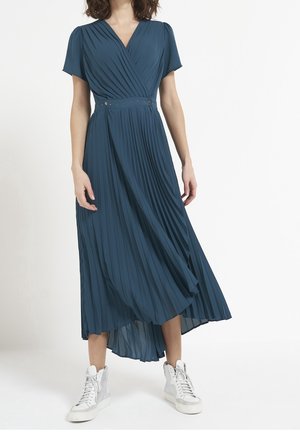 Mujer con vestido midi plisado de color verde azulado con mangas cortas y zapatillas blancas altas, de pie contra un fondo blanco.