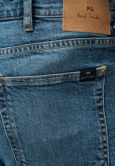 Τζιν παντελόνι από τζην με ξεθωριασμένο μπλε χρώμα, με αντίθετη πορτοκαλί ραφή. Δερμάτινο ταμπελάκι με την επιγραφή "PS Paul Smith" στη πίσω μέση.