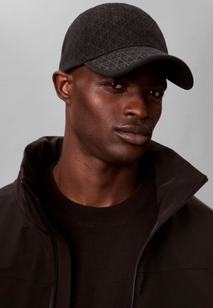 Homme portant une casquette de baseball noire à motifs et une veste noire zippée, regardant légèrement sur le côté devant un fond gris uni.