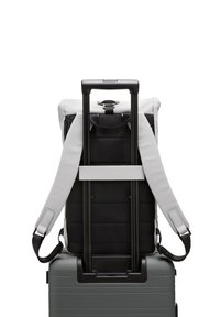 Sac à dos noir et blanc avec des bretelles rembourrées, attaché à une valise grise sur roulettes. Le sac à dos présente un fermoir et des détails de couture visibles.