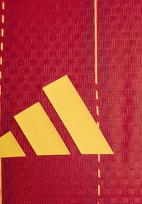 Un tissu rouge présente un motif texturé avec un logo Adidas jaune, composé de trois bandes diagonales et d'une ligne de couture jaune.