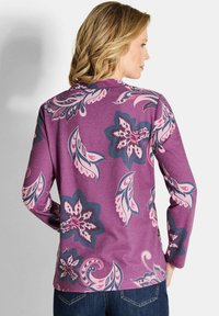 Vrouw met blond haar in een paars langmouwen shirt met grote roze en marineblauwe bloemenpatronen, gecombineerd met donkerblauwe jeans, zijwaarts gekeerd.