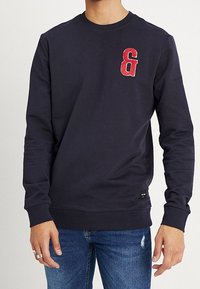 Granatowy sweter z okrągłym dekoltem, ozdobiony czerwonym haftowanym znakiem ampersand na lewej piersi, połączony z niebieskimi dżinsami.