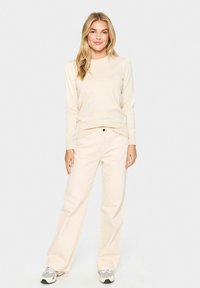 Bež srajca z dolgimi rokavi v kombinaciji s širokimi kremnimi hlačami. Obleka je narejena iz mehkega blaga, ima okroglo ovratnico in subtilno teksturo.