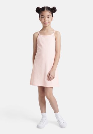 Ragazza giovane con due chignon che indossa un vestito senza maniche rosa pallido, calzini bianchi e scarpe da ginnastica bianche, in piedi su uno sfondo bianco.