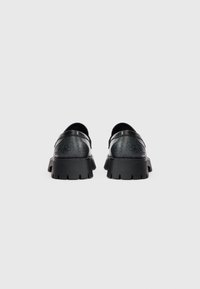 Mustat nahkaiset slip-on-kengät, joissa on teksturoitu design, paksut kumipohjat ja pyöristetyt varpaat. Ne ovat virtaviivaisia ja yksinkertaisia ilman ylimääräisiä koristeita.