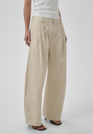 Nő bézs széles szárú nadrágban és fehér ujjatlan felsőben, fekete, nyitott orrú, sarkú szandálban áll egy egyszínű háttér előtt.