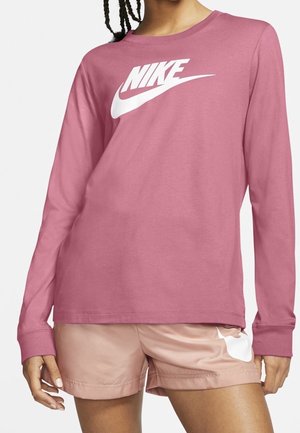 Person trägt ein staubrosafarbenes langärmeliges Nike-Shirt mit weißem Logo und hellrosa Shorts, die teilweise den Nike-Swoosh zeigen.