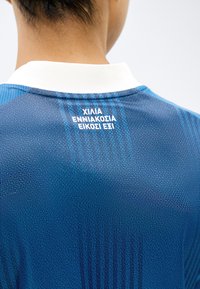 Άτομο που φοράει μπλε αθλητική φανέλα με άσπρο γιακά, στην πίσω πάνω πλευρά εμφανίζεται το ελληνικό κείμενο "ΧΙΛΙΑ ΕΝΝΙΑΚΟΣΙΑ ΕΙΚΟΣΙ ΕΞΙ".