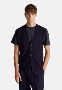 Chaleco de punto azul marino con diseño de cuello en V, botones delanteros y textura suave, usado sobre una camiseta gris oscuro. Corte ajustado.