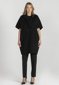 Czarna wełniana odzież wierzchnia o luźnym kroju, z dużymi klapami, podwójnymi guzikami i przednimi kieszeniami, w zestawieniu z obcisłymi czarnymi spodniami i wysokimi obcasami.