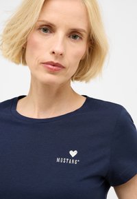 Námořnická modrá bavlněná tričko s kulatým výstřihem a krátkým rukávem; má na hrudi bílé vyšívané srdce a logo "MUSTANG".