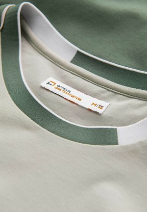 Světlé zelené tričko s tmavším zeleným pruhovaným límcem a bílým lemem. Štítek uvádí "ombre performance," velikost M. Měkký bavlněný materiál.