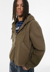 Oliven grøn zip-hoodie med hætte, kengurulommer og ribbede manchetter, lavet af blødt stof. Har syninger på brystet.
