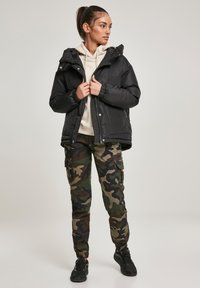 Čierna prešívaná bunda s kapucňou, zapínaním na zips a elastickými manžetami, nosená cez krémovú mikinu s kapucňou. Kombinovaná s maskáčovými cargo nohavicami a čiernymi teniskami.