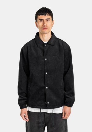 Mann trägt eine schwarze, geknöpfte Jacke mit elastischen Bündchen, darunter ein weißes Hemd und dunkle Hose, steht vor einem einfarbigen hellen Hintergrund.