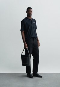 Fekete poloing gallérral és logóval, fekete nadrággal és bőr cipővel párosítva. Egy struktúrált fekete táskát tart, arany részletekkel.