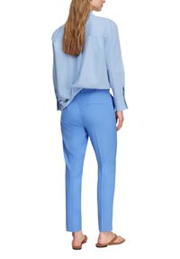 Kobieta w jasnoniebieskiej koszuli z długim rękawem i dopasowanych, wąskich spodniach, stojąca boso na białym tle.