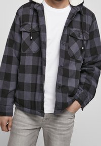 Svart och grått flanelljacka med en Sherpa-fodrad huva, knäppningar, bröstfickor och en avslappnad passform. Bärs över en vit t-shirt.