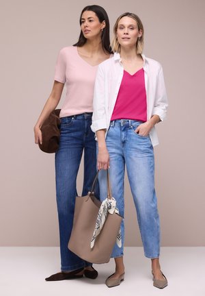 Dvě ženy stojí vedle sebe; jedna má růžový top s tmavými džínami a drží hnědé psaníčko, druhá bílou košili přes jasně růžový top s světlými džínami a drží béžovou tašku přes rameno.