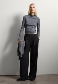 Maglietta grigia a maniche lunghe con silhouette aderente, abbinata a pantaloni neri larghi e a vita alta; tiene un maglione grigio abbinato.