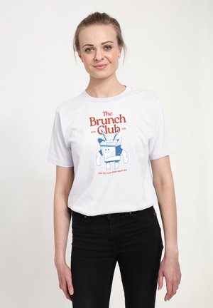 Person iført en hvid T-shirt med teksten “The Brunch Club” og en tegneserie-vaffel med bestik, stående mod en ensfarvet baggrund.