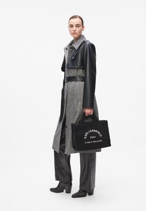 Grijze wollen mix jas met leren accenten, een ceintuur in de taille en een grote kraag. Zwarte rechthoekige tas met witte tekst. Zwarte op maat gemaakte broek en haklaarzen.