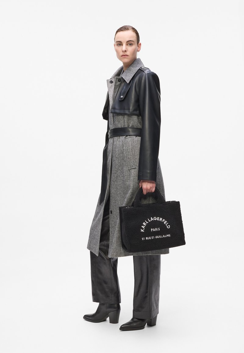 Grijze wollen mix jas met leren accenten, een ceintuur in de taille en een grote kraag. Zwarte rechthoekige tas met witte tekst. Zwarte op maat gemaakte broek en haklaarzen.