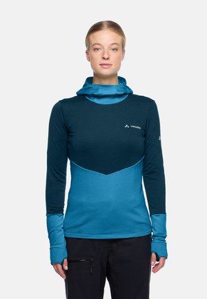 Vrouw in een twee-kleurige blauwe lange mouw sporttop met duimgaten en een ingebouwde nekkraag, staand tegen een effen witte achtergrond.