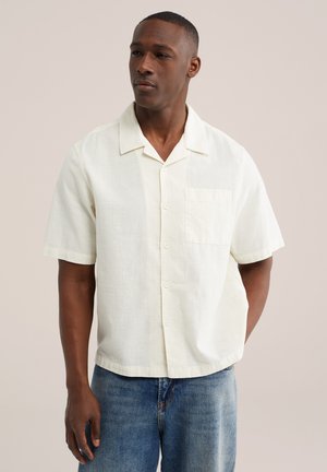 Muž v krátkym rukávom bielom zapínacom tričku s náprsným vreckom a modrých džínsoch, stojaci pred jednofarebným pozadím.