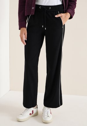 Pantalon large noir avec des bandes blanches sur les côtés, taille élastique et cordon de serrage. Associé à des baskets blanches et un pull texturé bordeaux.