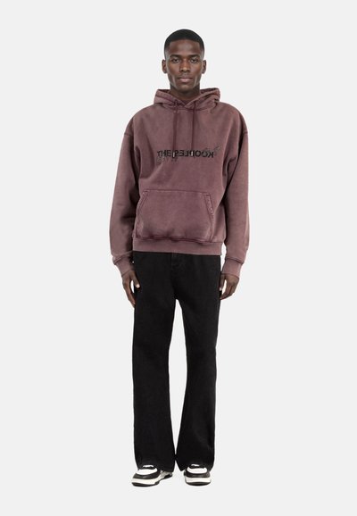Bruine hoodie met een voorzak en contrasterende zwarte tekst, gecombineerd met zwarte corduroy broek en zwart-witte sneakers.