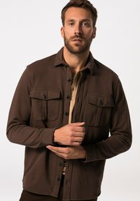 Bruine button-up jas met twee borstzakken, zachte stof, lange mouwen en een kraag. Gedragen over een beige shirt.