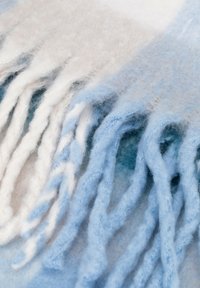 Wełniany szalik z miękkimi frędzlami na brzegach, w odcieniach jasnoniebieskiego i białego, z gradientowym wzorem i miękką, puszystą fakturą.