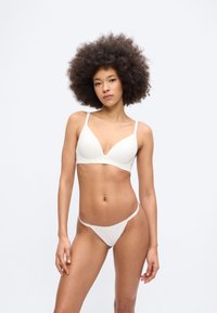 Γυναίκα με μεγάλο σγουρό μαλλί που φοράει λευκό σουτιέν και εσώρουχα που ταιριάζουν, στέκεται μπροστά σε απλό φωτεινό φόντο με ουδέτερη έκφραση.