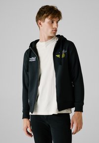 Crna zip-up dukserica s kapuljačom na vezanje, s Red Bull Racing logotipima na prsima i gornjem dijelu rukava, nosi se preko svijetle košulje.