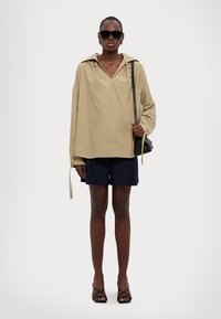 Camicia beige oversize con colletto a punta, abbinata a pantaloni shorts blu navy. L'outfit è completato da sandali neri con cinturini e occhiali da sole.