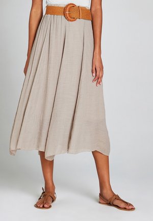 Vrouw met een beige midi-rok met plooien, een brede gevlochten tan riem en tan platte sandalen tegen een effen achtergrond.