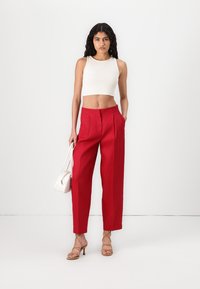 Pantalons taille haute rouges avec plis, en tissu léger. Associés à un crop top blanc et des sandales à lanières beiges, tenant un sac blanc.